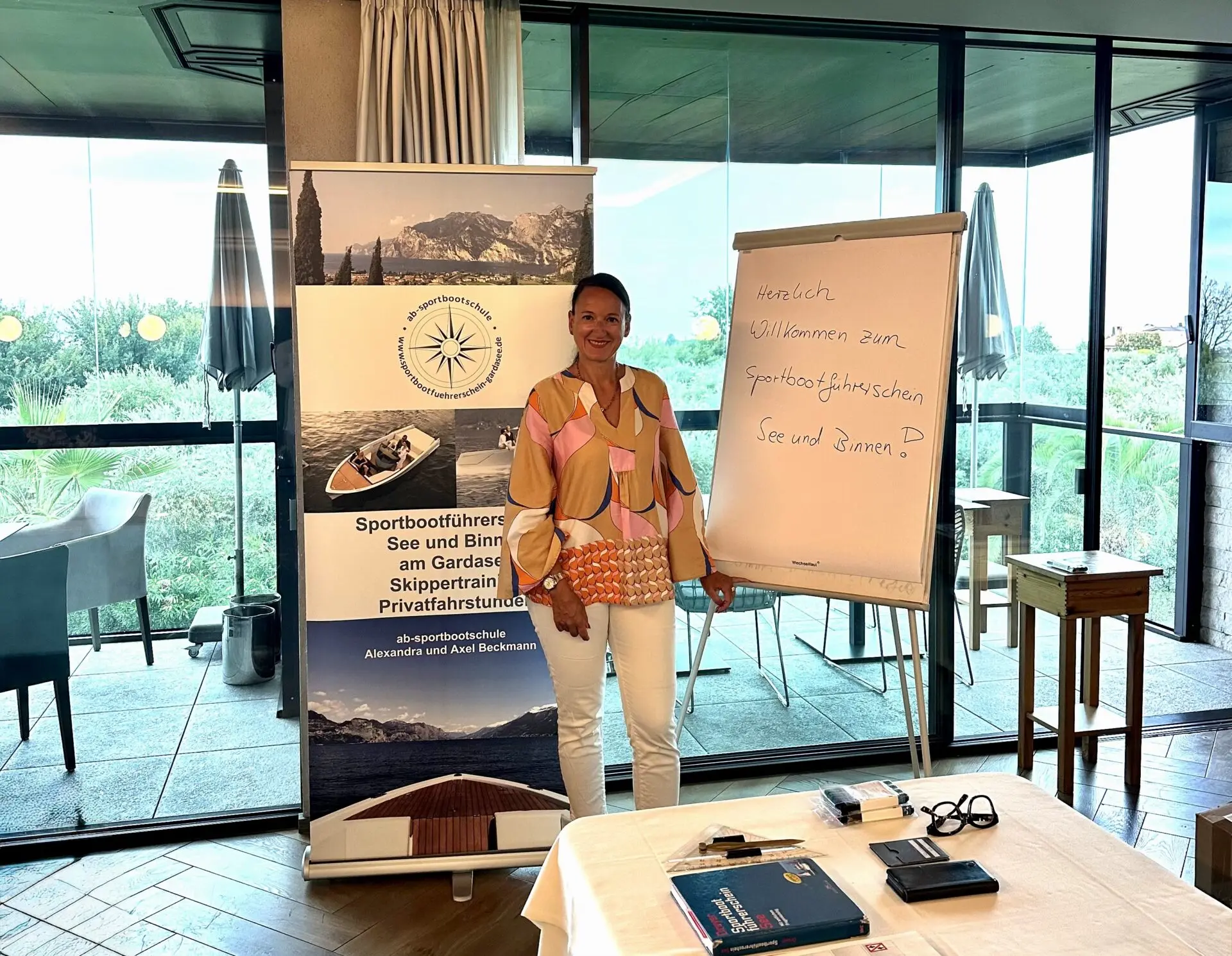 Frau steht neben einem Flipchart und einem Banner der Sportbootführerscheine am Gardasee mit Informationen zu Kursen und Skippern.