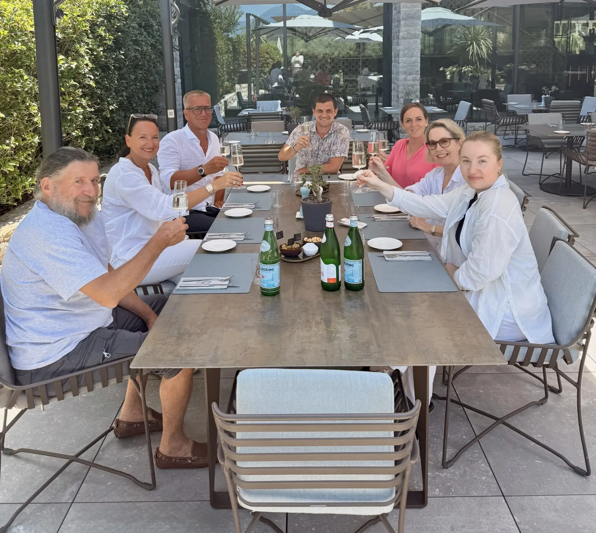 Gruppe von sieben Personen, die an einem Tisch im Freien auf einer Terrasse sitzen und mit Gläsern anstoßen.