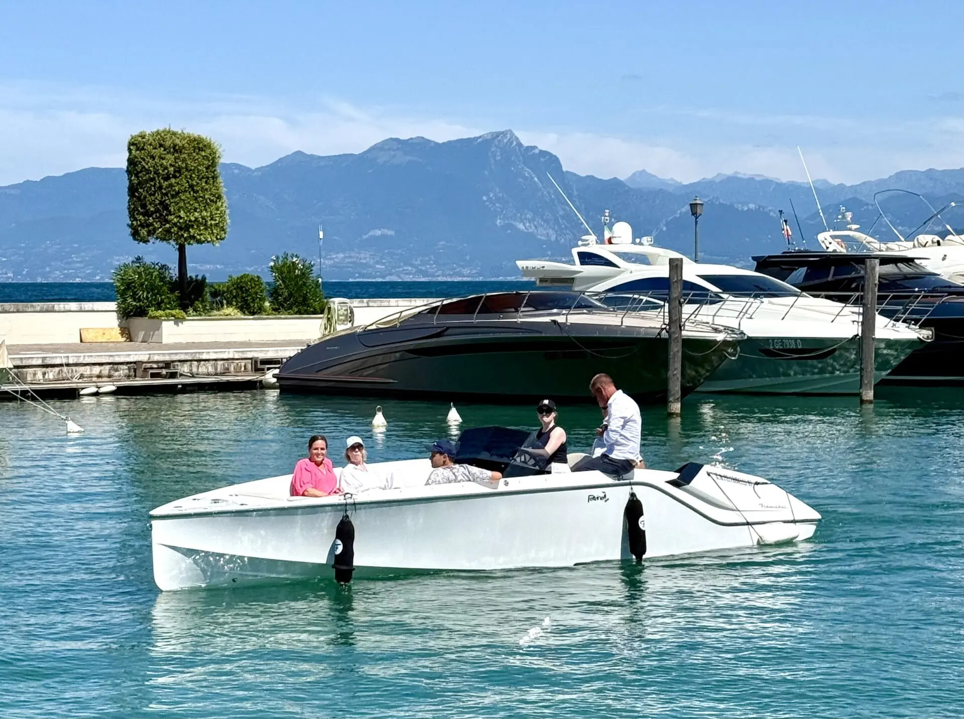 Ein modernes Boot mit mehreren Personen, das auf dem Wasser im Hafen von Lazise am Gardasee fährt, umgeben von anderen Yachten.