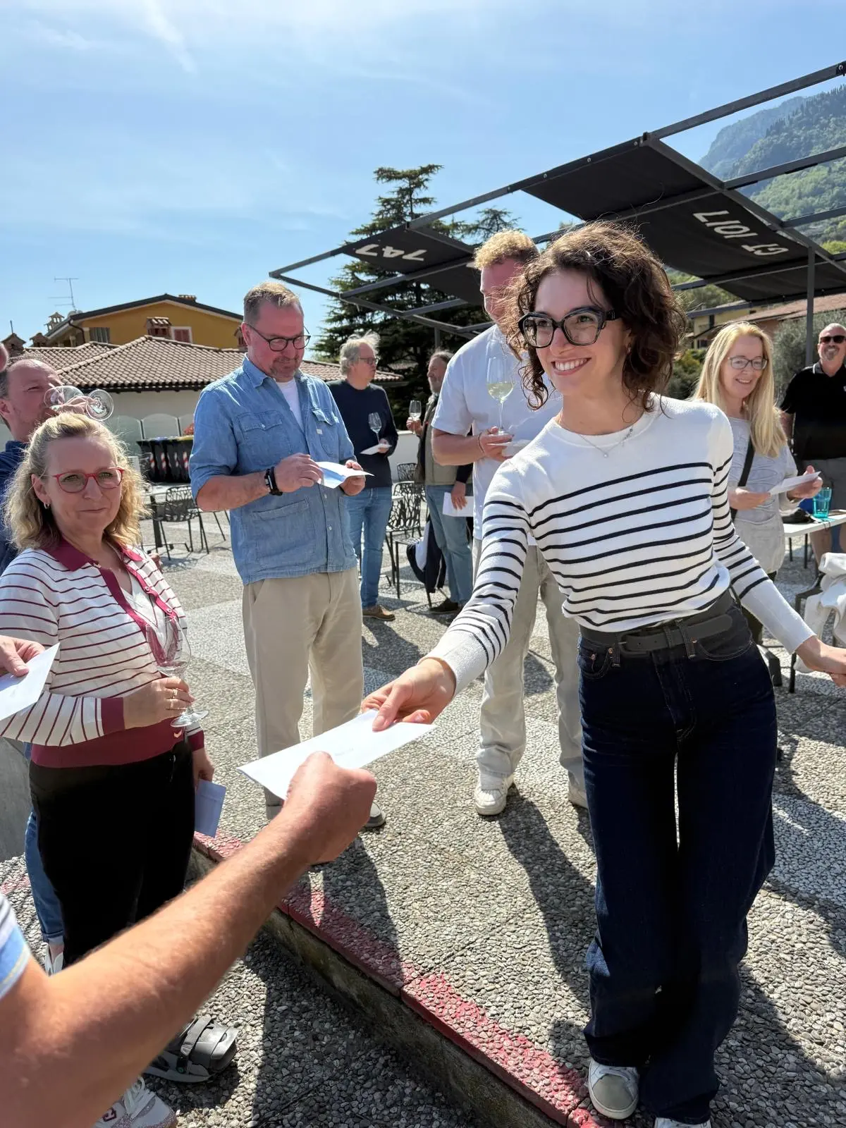 Feierliche Stimmung am Gardasee: Gruppe stößt draußen mit Gläsern an, gestreifte Oberteile, schwarze Überdachung und sonniges Ambiente in Gargnano.