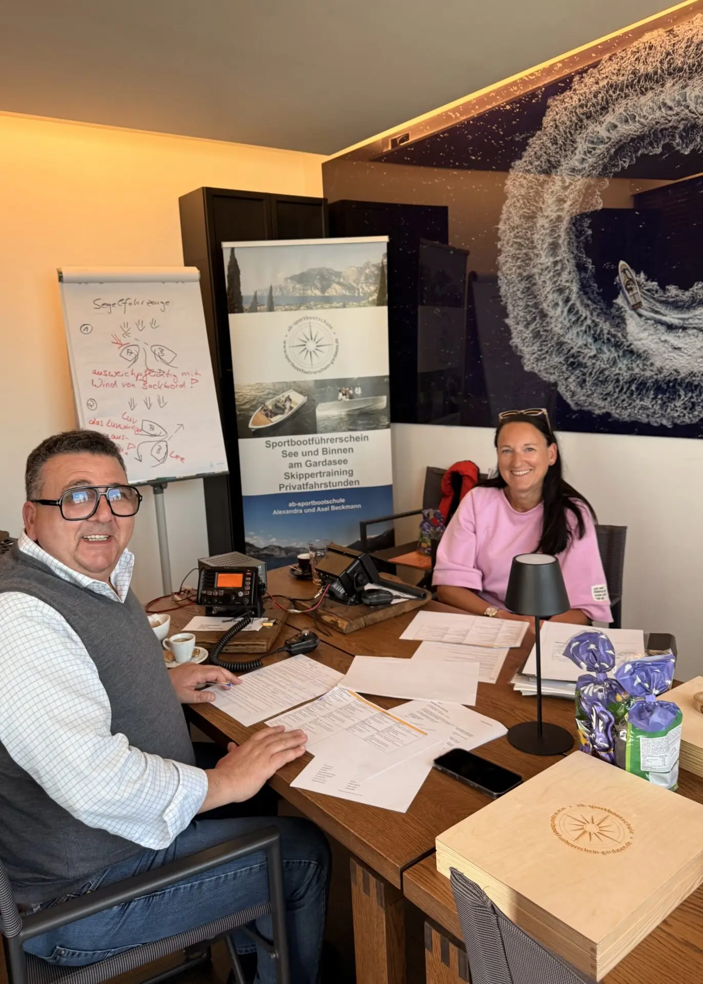 Im Schulungsraum mit Holztisch und weißen Unterlagen: Poster „Sportbootführerschein See und Binnen am Gardasee“, weißes Whiteboard, pinkes Oberteil