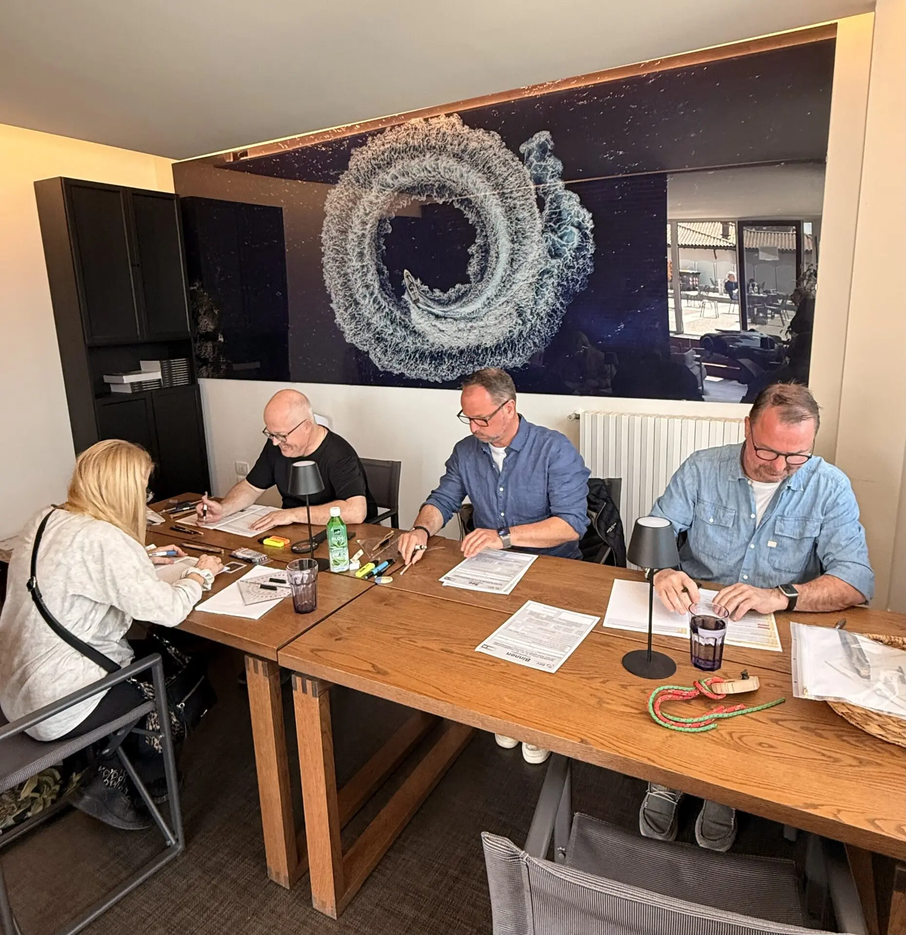 Bootsführerschein Gardasee Prüfung am Holztisch: mehrere Teilnehmer mit Brille und Unterlagen im Raum, im Stil des Clubhaus Feltrinellis, ruhig und konzentriert.