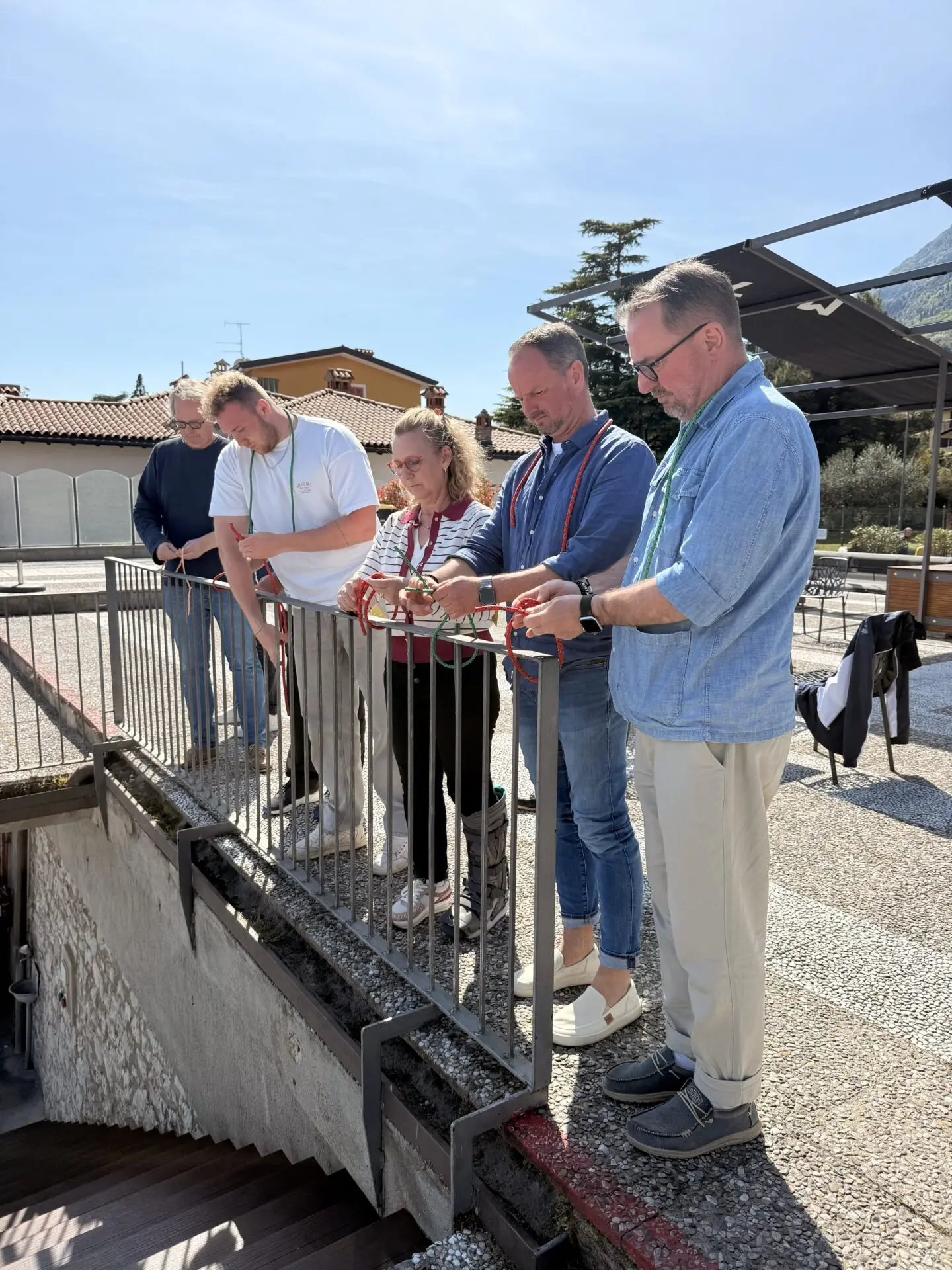 Gruppe bei Outdoor-Übung am Gardasee, Teilnehmer mit blauen Hemden und Jeans, Blick auf Geländer und Terrasse, sonniges Wetter.