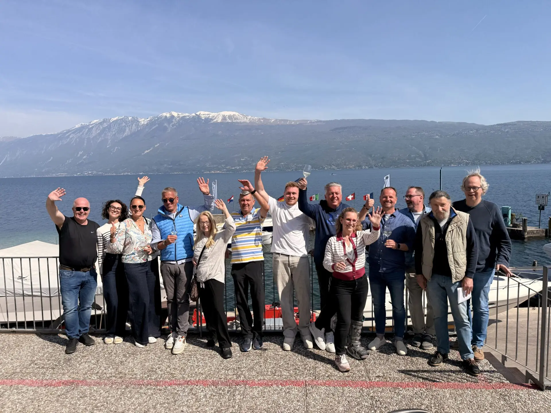 Teilnehmende am Ufer des Gardasees mit erhobenen Händen und Flaggen, im Hintergrund See und Alpengebirge bei klarem Himmel.