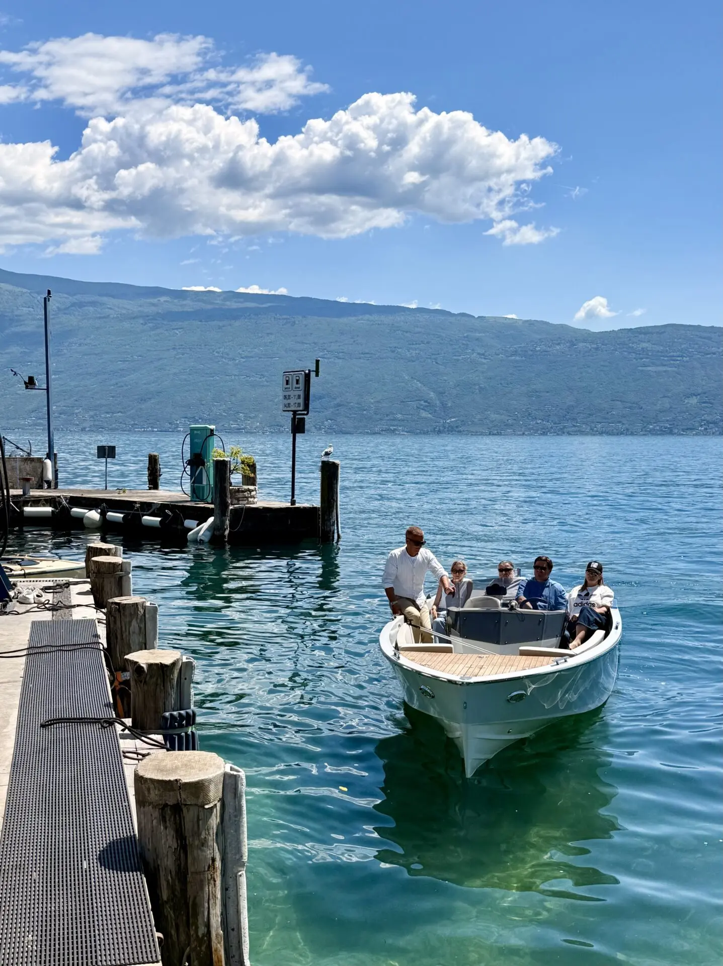 Boot mit Fahrgästen am Steg in Gargnano am Gardasee, vor Bergen, blauem Himmel und ruhigem Wasser bei der Nautica Feltrinelli.