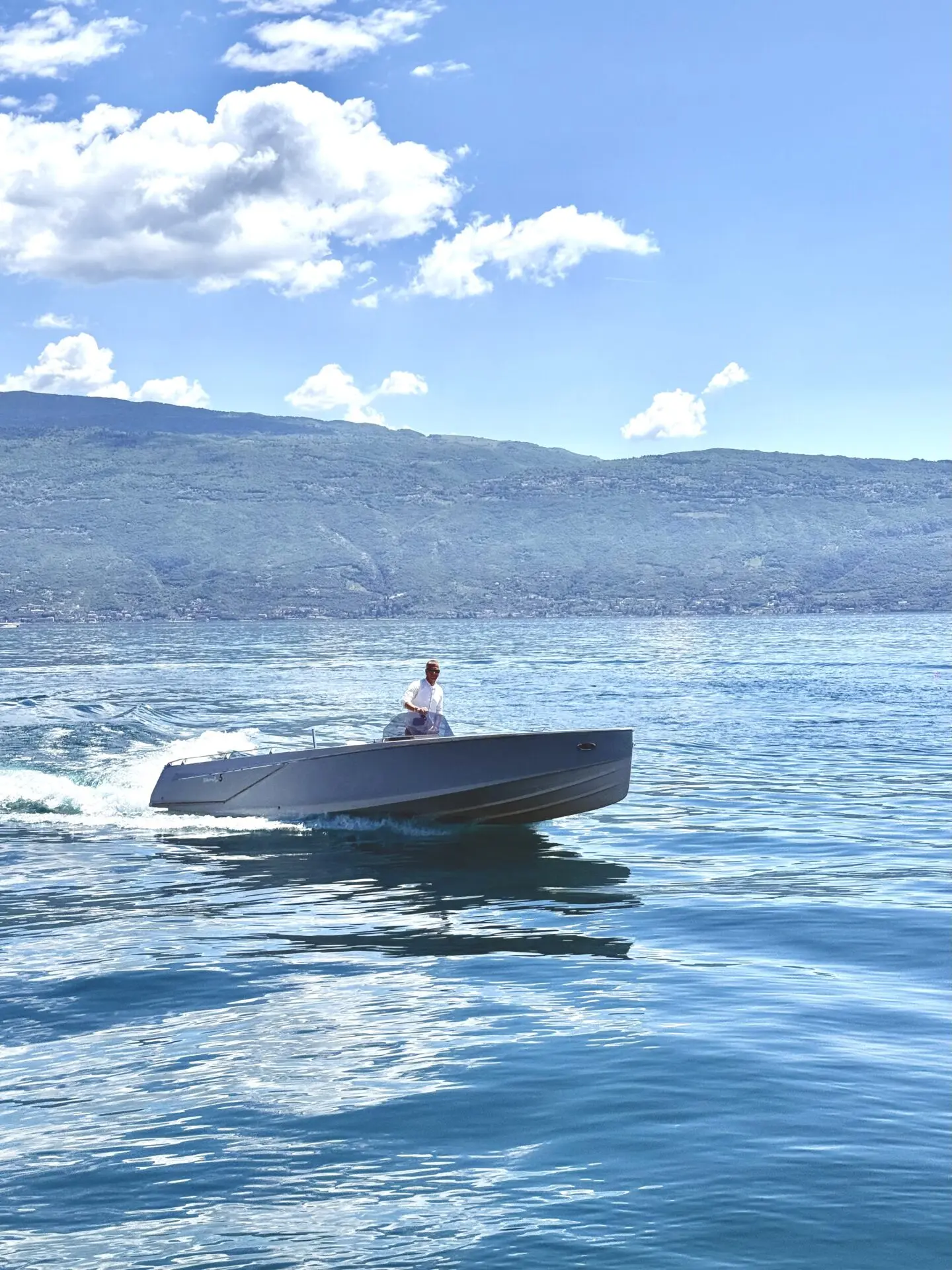 Auf dem Gardasee bei Gargnano fährt ein graues Motorboot auf ruhigem, blauem Wasser unter sonnigem Himmel vor der Bergkulisse.