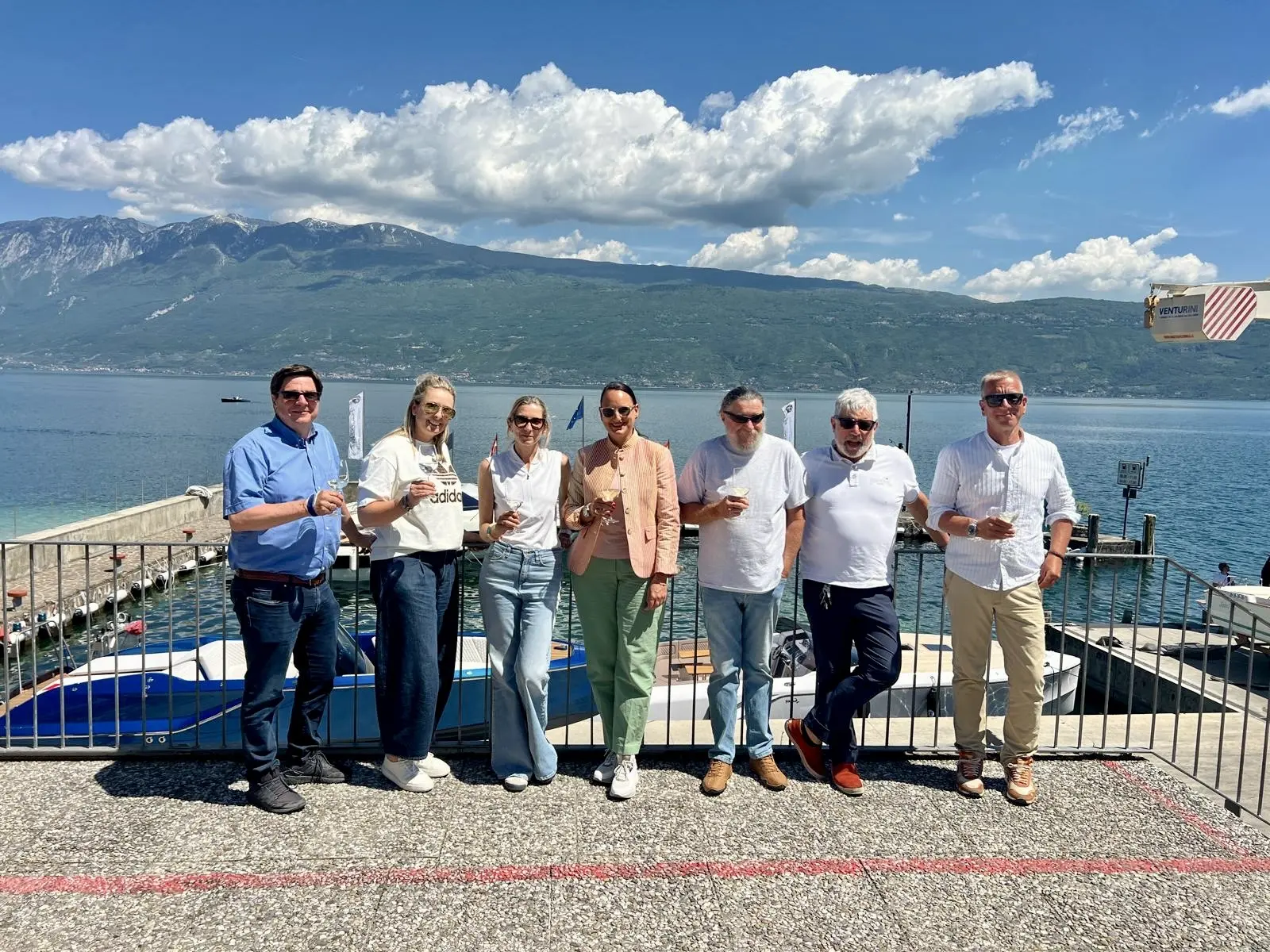 Vor dem Hafen in Gargnano am Gardasee steht eine Gruppe mit Getränken, dahinter liegen Frauscher Boote, Berge und ein weiter blauer Himmel.