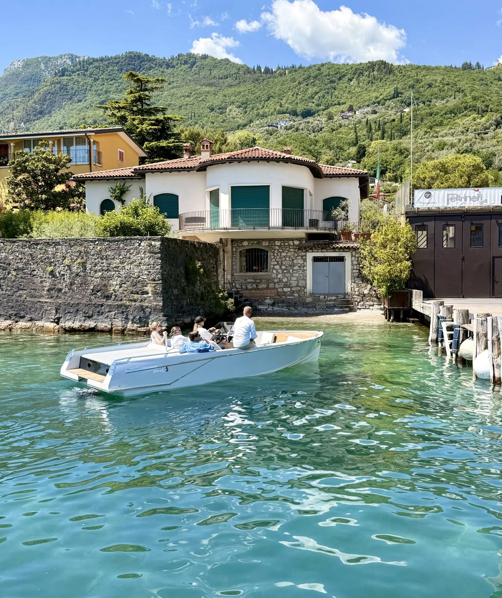 Auf dem Gardasee liegt ein weißes Motorboot vor Gargnano, im Hintergrund der Cantiere Nautico Feltrinelli und grüne Berghänge bei sonnigem Wetter.
